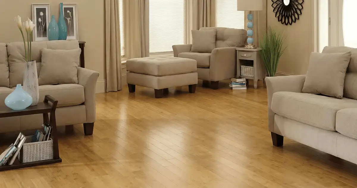 A living room with professionally installed bamboo flooring, beige sofas, a matching ottoman, a side table with a blue lamp, vases, framed pictures, and large windows with blinds and curtains in Lakeland, FL